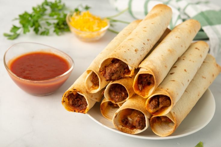 taquitos stacked on a plate with salsa by it