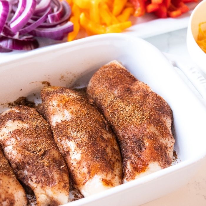 seasoned chicken in casserole pan with veggies behind it