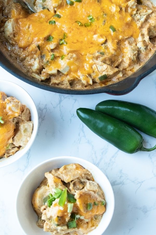bowls of green chili chicken with pan behind it and peppers on marble counter
